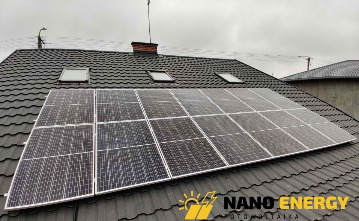 Panel słoneczny na ciemnym dachu z dachówką, widoczne okna dachowe i komin z cegły, pochmurne niebo w tle.