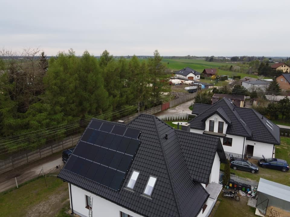 Widok z góry na dom jednorodzinny z panelami słonecznymi na dachu, w tle zabudowa wiejska i drzewa, pochmurne niebo.