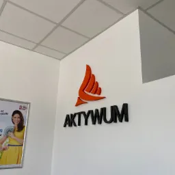 Logo firmy 'AKTYWUM' z pomarańczowym symbolem graficznym, umieszczone na białej ścianie w biurze, obok reklama z uśmiechniętą kobietą trzymającą banknoty.