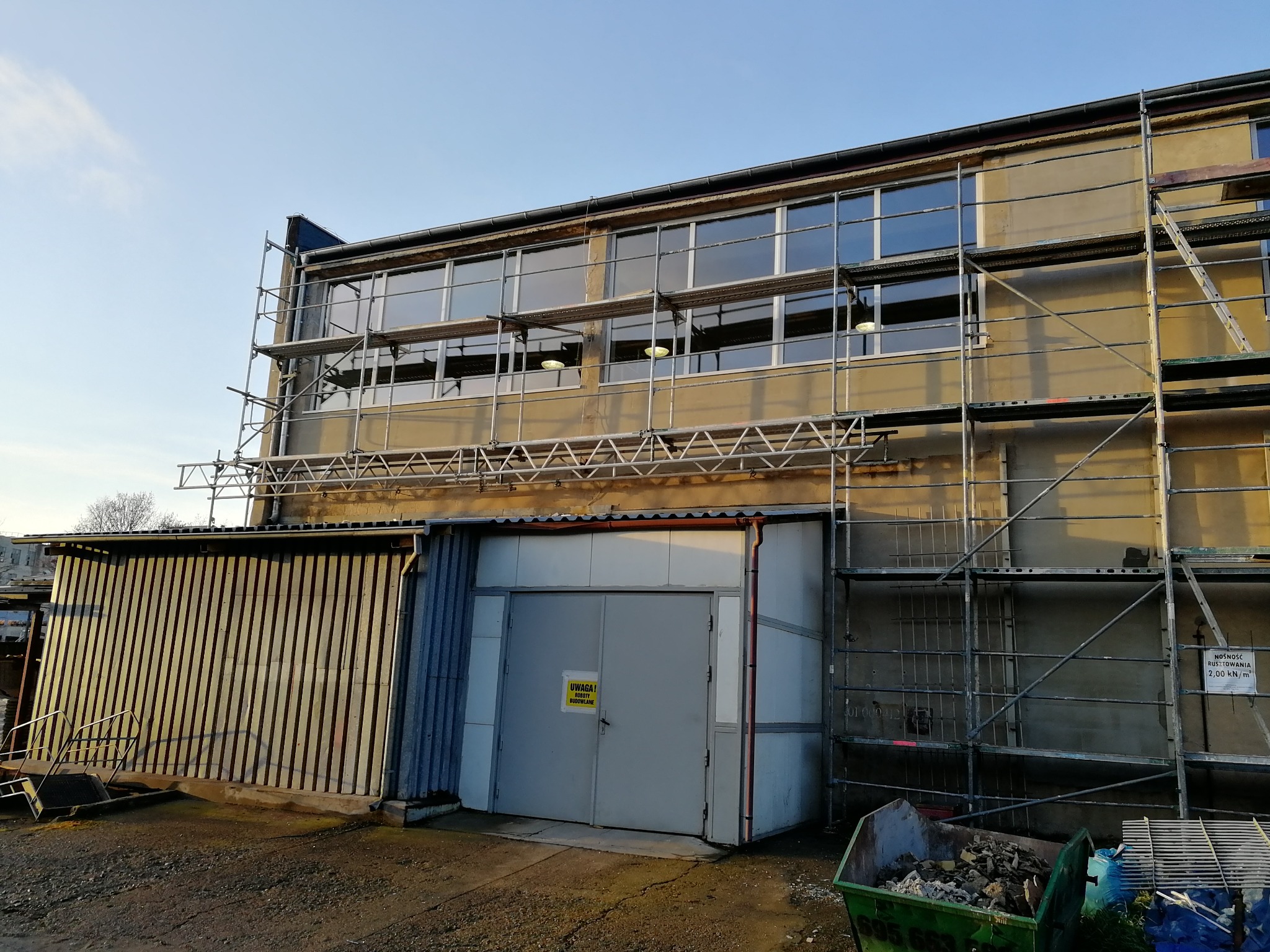 Budynek przemysłowy w trakcie remontu, z rusztowaniem, nowymi oknami i kontenerem na gruz na pierwszym planie.