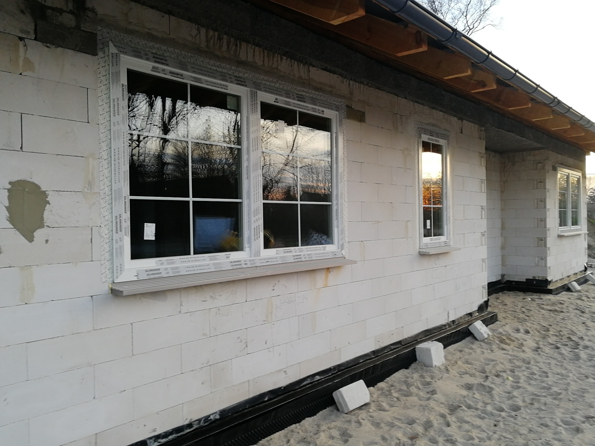 Nowe białe okna z kratownicą w stanie surowym budynku z białych bloczków, widoczne zabezpieczenia na ramach okiennych, piasek przed budynkiem.