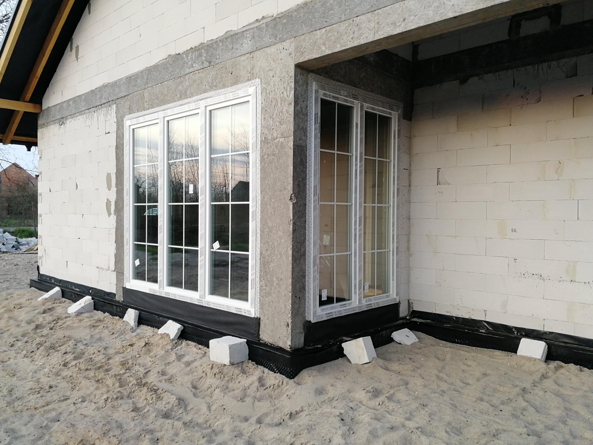 Dwa białe okna z białymi szprosami, zamontowane w surowym stanie budynku z białego bloczka, zabezpieczone czarną folią hydroizolacyjną i podparte białymi bloczkami na piaszczystym podłożu.