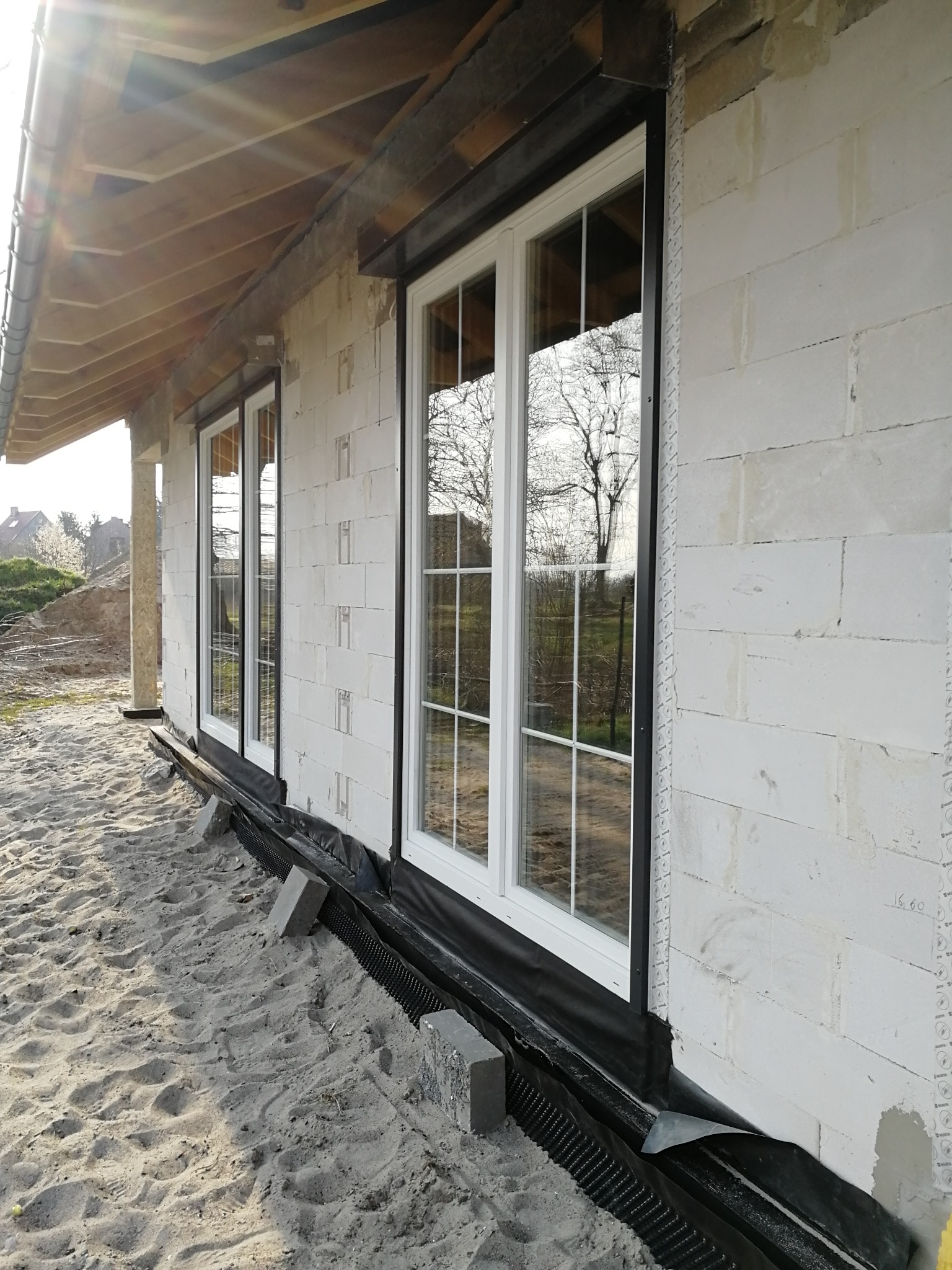 Montaż białych okien z czarnymi roletami zewnętrznymi w surowym budynku z białego bloczka, widoczna izolacja fundamentów i piasek.