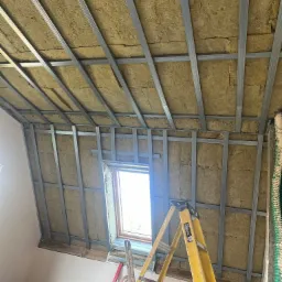 Montaż metalowego rusztu pod płyty gipsowo-kartonowe na poddaszu w Oławie, z widoczną izolacją z wełny mineralnej i oknem dachowym.