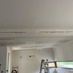 BHN-BUD - Wnętrze w trakcie prac wykończeniowych: zamontowane płyty gipsowo-kartonowe na suficie i ścianach, widoczne szpachlowane łączenia, drabina Krause i drewniana drabinka w pomieszczeniu z oknem.