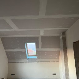 BHN-BUD - Poddasze z oknem dachowym w trakcie wykańczania płytami gipsowo-kartonowymi, widoczne łączenia płyt wymagające szpachlowania, instalacja elektryczna wystaje ze ściany.