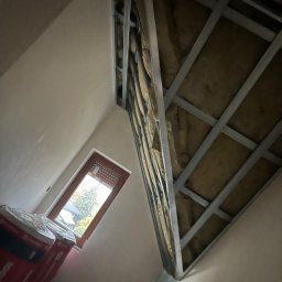 BHN-BUD - Skośne poddasze w trakcie ocieplania wełną mineralną, widoczne metalowe profile konstrukcyjne i częściowo zakryte rolki izolacji przy oknie.