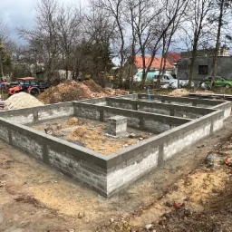 Fundamenty domu jednorodzinnego z bloczków betonowych, częściowo zalane betonem, na placu budowy z widocznymi maszynami rolniczymi w tle.