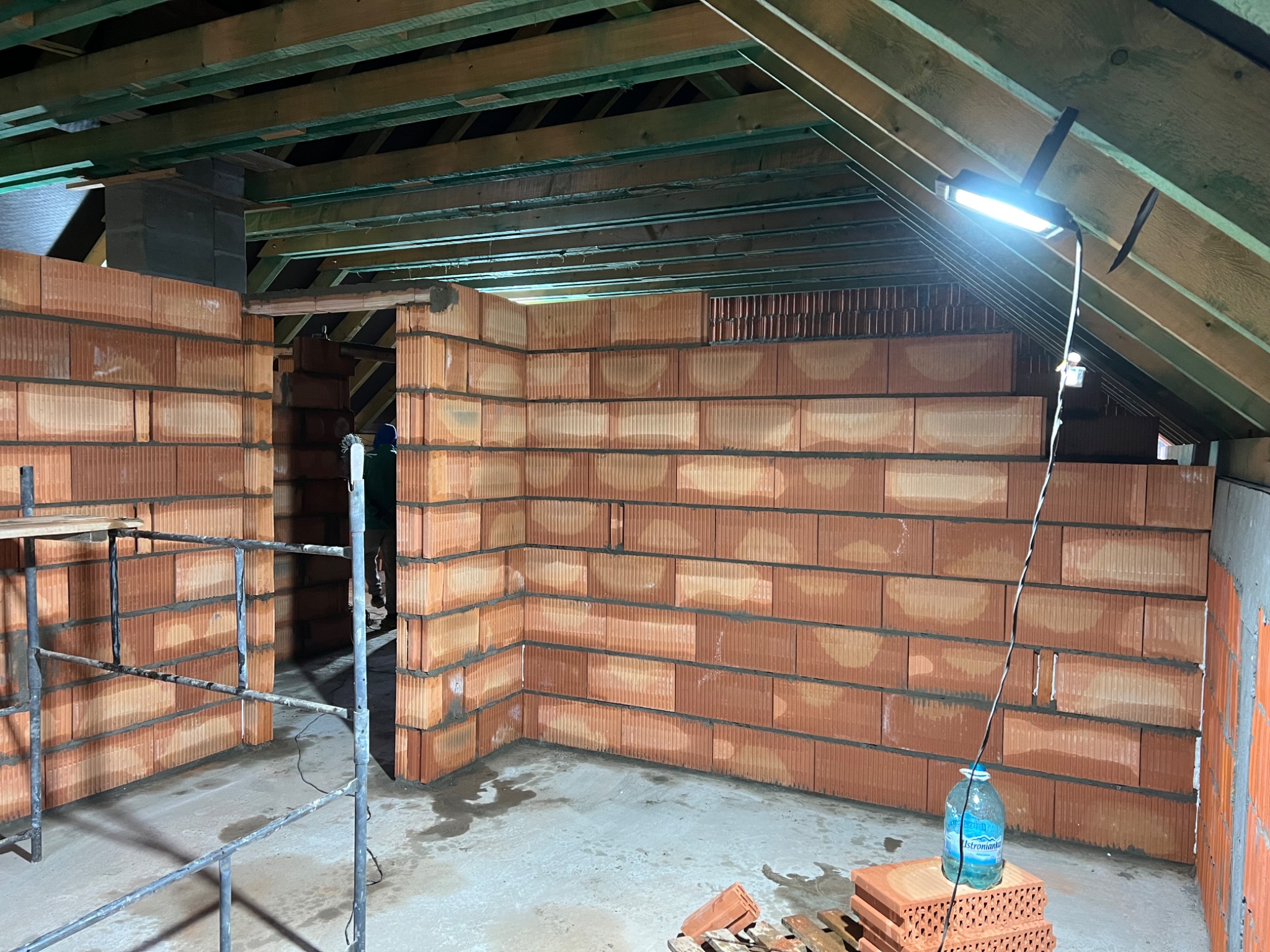 Wnętrze domu w Oławie w trakcie budowy z czerwonej cegły, widoczne ściany działowe, drewniana konstrukcja dachu, lampa budowlana i rusztowanie.