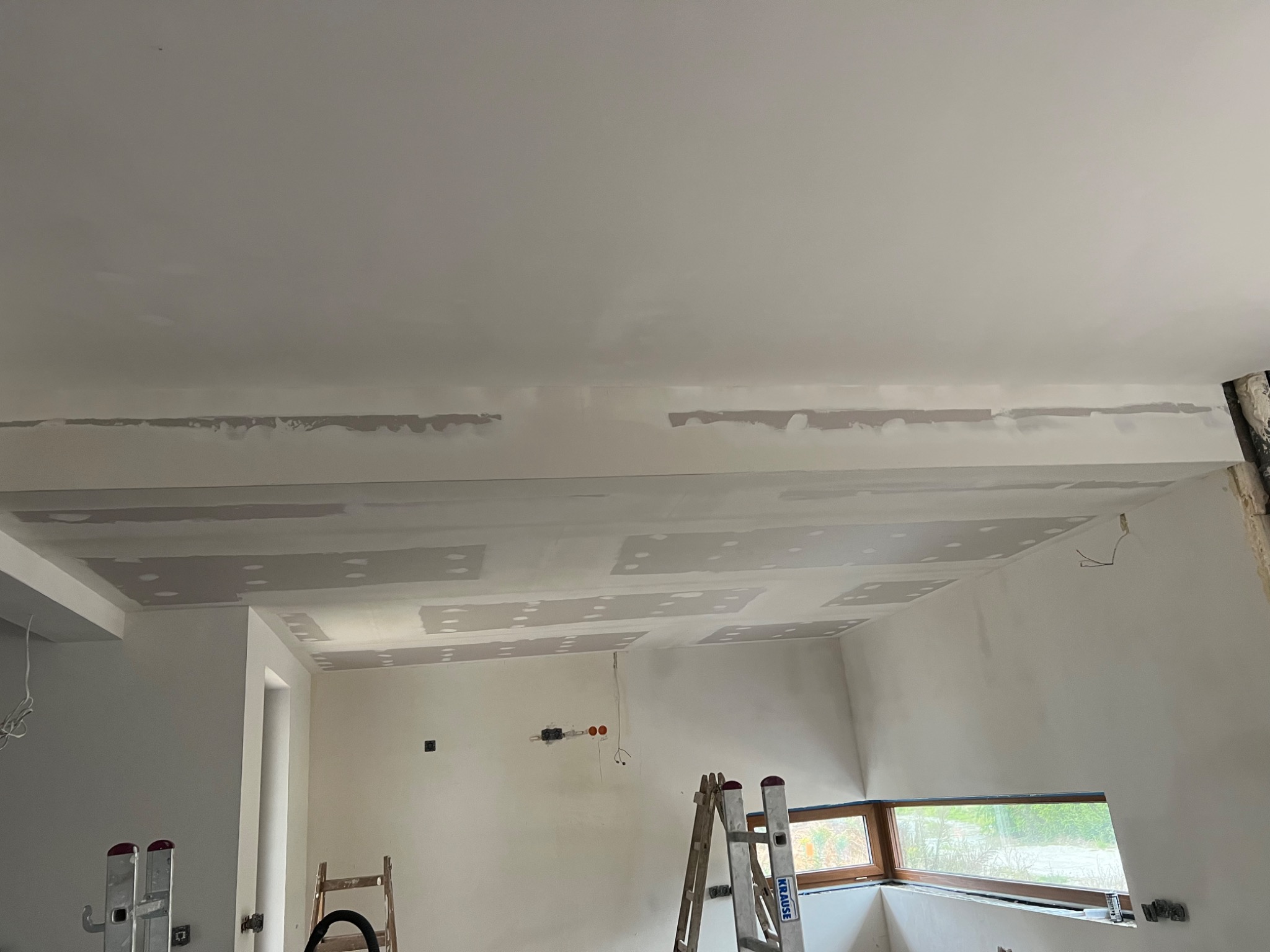 Wnętrze w trakcie prac wykończeniowych: zamontowane płyty gipsowo-kartonowe na suficie i ścianach, widoczne szpachlowane łączenia, drabina Krause i drewniana drabinka w pomieszczeniu z oknem.