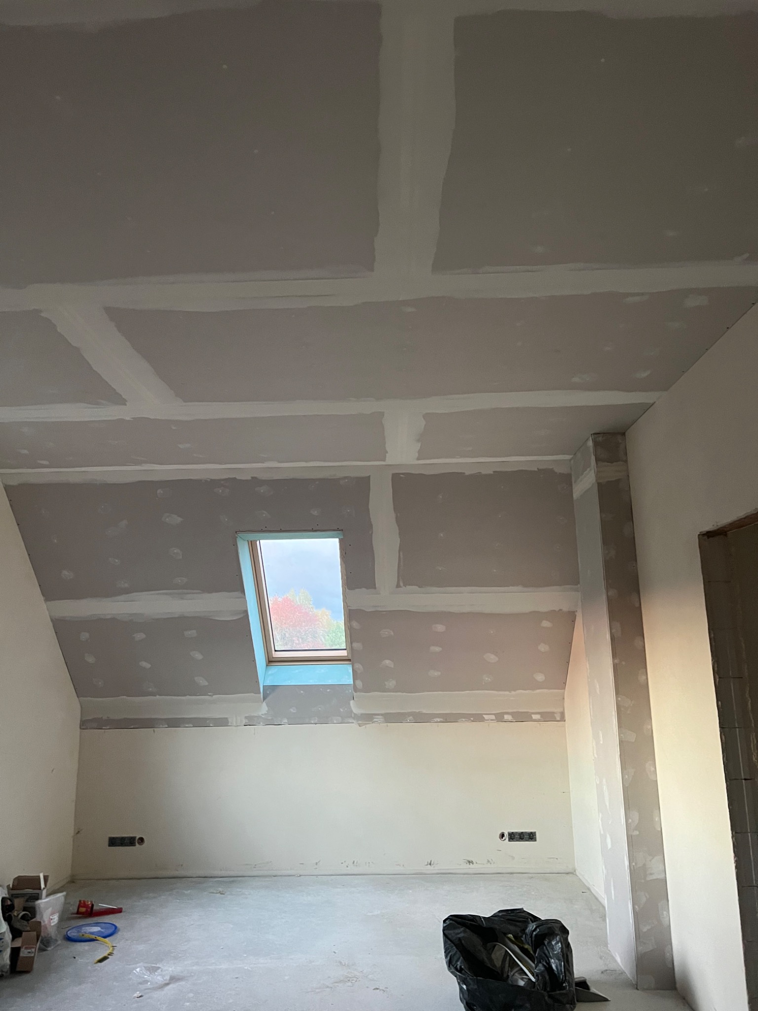 Poddasze z oknem dachowym w trakcie wykańczania płytami gipsowo-kartonowymi, widoczne łączenia płyt wymagające szpachlowania, instalacja elektryczna wystaje ze ściany.