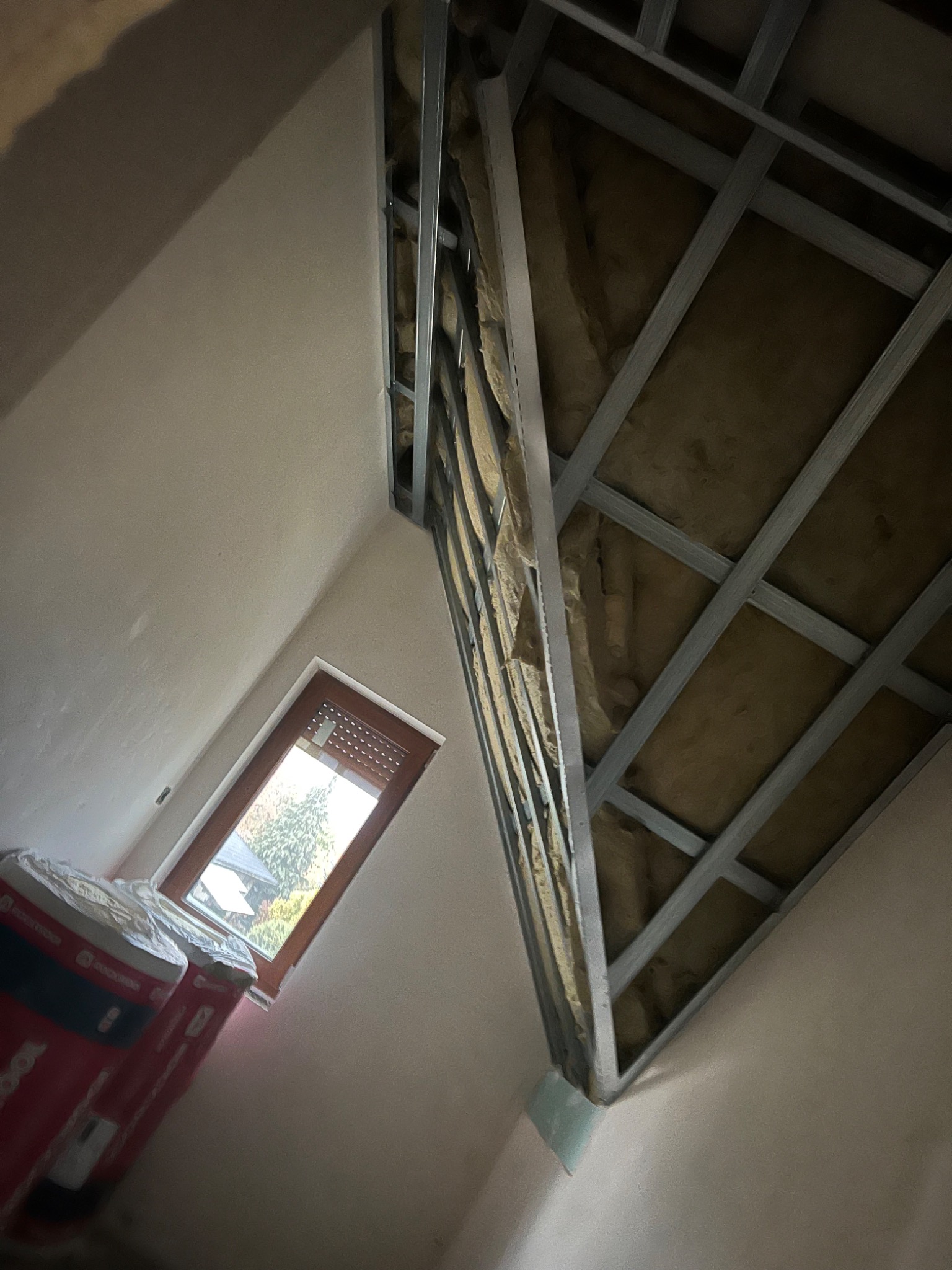 Skośne poddasze w trakcie ocieplania wełną mineralną, widoczne metalowe profile konstrukcyjne i częściowo zakryte rolki izolacji przy oknie.
