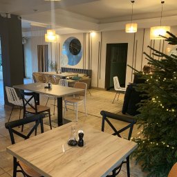 Oliwia Kalandyk Architektura Wnętrz - Jasne wnętrze restauracji z drewnianymi stołami, rattanowymi krzesłami, dekoracyjnym panelem ściennym z motywem marmuru i choinką w rogu.