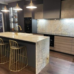Oliwia Kalandyk Architektura Wnętrz - Nowoczesna kuchnia z wyspą, blatem z jasnego granitu, złotą armaturą i hokerami oraz podświetlaną szafką z alkoholem w tle.