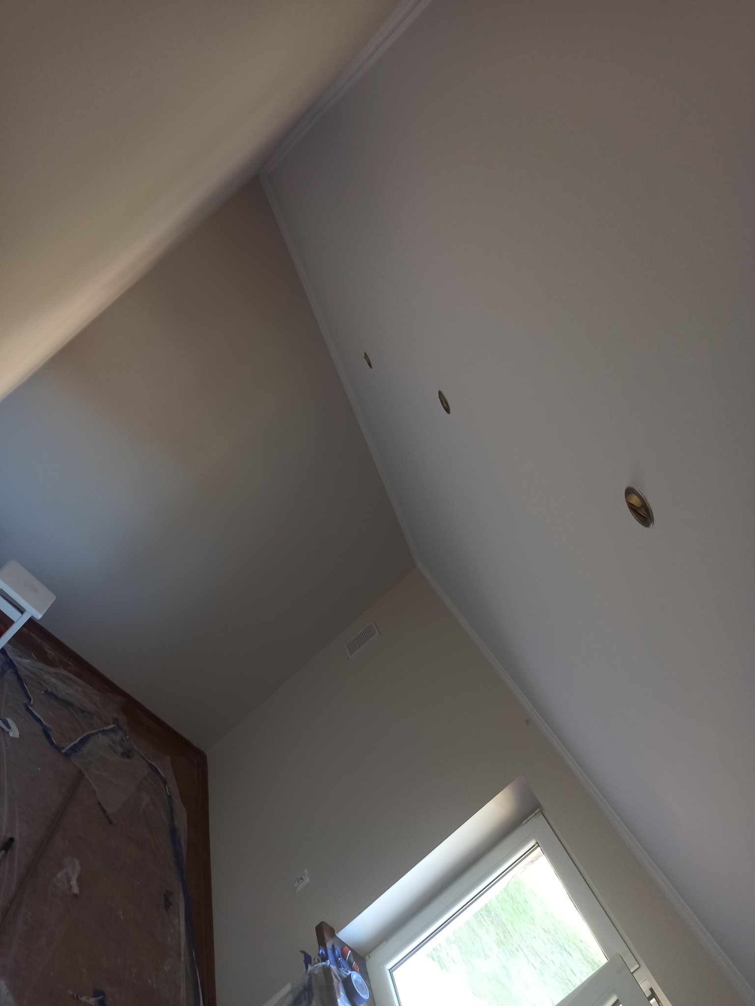 Skośny sufit po malowaniu na biało z trzema punktowymi otworami na oświetlenie, widoczne listwy przypodłogowe, okno i zabezpieczona folią podłoga.