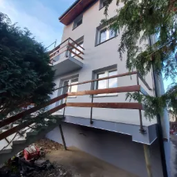 Dwupiętrowy dom z szarą elewacją i drewnianymi barierkami balkonowymi, widok z dołu z elementami roślinności na pierwszym planie i niebieskim niebem w tle.