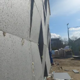 Szara elewacja budynku z dekoracyjnymi, geometrycznymi wcięciami, w trakcie ocieplania, widoczne płyty izolacyjne i pianka montażowa, na ziemi materiały budowlane i śmieci.