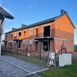 Dachy Kowalski Andrzej Kowalski - Surowy stan szeregowca z cegły, z czarną dachówką i kominami, w trakcie budowy. Widoczny pracownik na dachu. Ujęcie z zewnątrz, w słoneczny dzień.