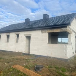 Dachy Kowalski Andrzej Kowalski - Niedokończony dom z ciemnym dachem z blachy na tle błękitnego nieba. Widoczne kominy i elementy konstrukcyjne. Na pierwszym planie ziemia i resztki materiałów budowlanych.