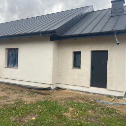 Dachy Kowalski Andrzej Kowalski - Nowoczesny dom jednorodzinny z ciemnoszarą blachodachówką i świeżo otynkowaną elewacją. Okna zabezpieczone folią, widoczny fragment ogrodu i niebieski wąż.