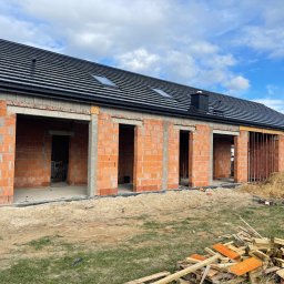 Dachy Kowalski Andrzej Kowalski - Dom w trakcie budowy z czarnym dachem i ceglanymi ścianami. Widoczne okna dachowe i komin. Na pierwszym planie sterta drewna i piasku.