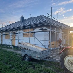 Dachy Kowalski Andrzej Kowalski - Dom w trakcie budowy z rusztowaniem i przyczepką. Widoczny fragment traktora. Dach pokryty szarą dachówką. Prace budowlane w toku, ściany ocieplone styropianem.