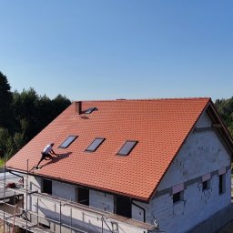 Dachy Kowalski Andrzej Kowalski - Czerwony dach z lukarnami w Wołczynie, pokryty dachówką karpiówką. Dekarz pracuje na dachu, widoczne rusztowania i surowy stan budynku. Słoneczny dzień.