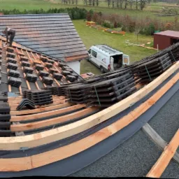 Panoramiczne ujęcie dachu w trakcie krycia dachówką ceramiczną, widoczna konstrukcja więźby, membrana dachowa oraz ułożone w stosy dachówki, w tle zabudowa jednorodzinna i zieleń.