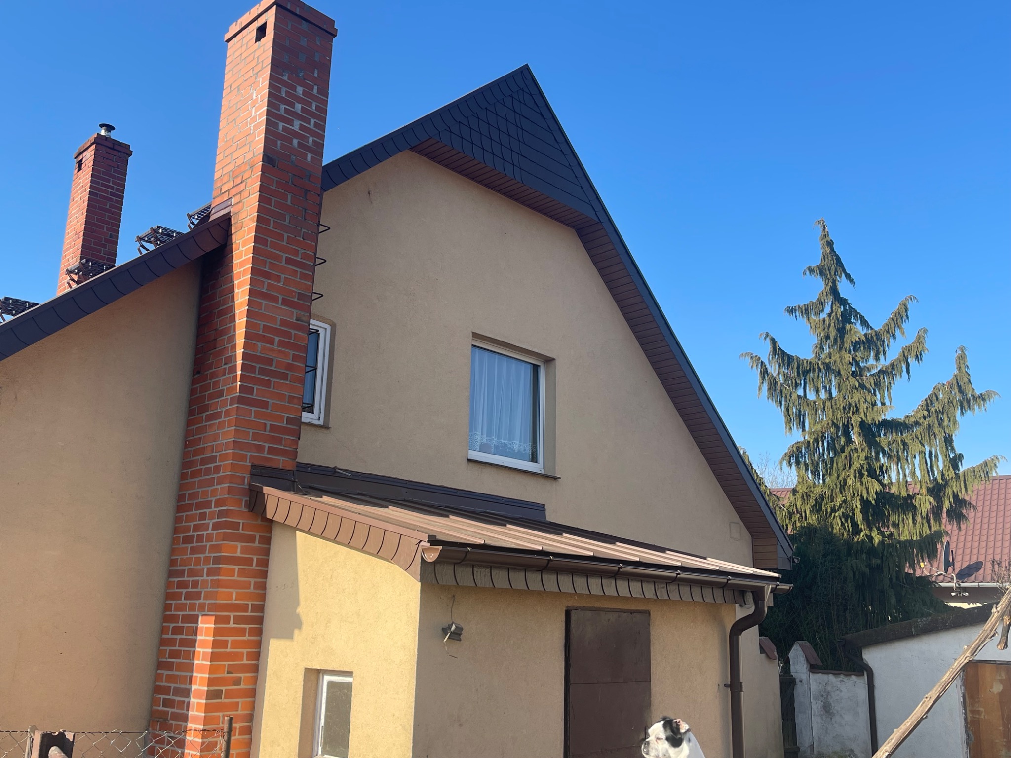 Dom jednorodzinny z ciemną dachówką i ceglanym kominem na tle błękitnego nieba. Widoczny fragment elewacji i iglaste drzewo w tle.