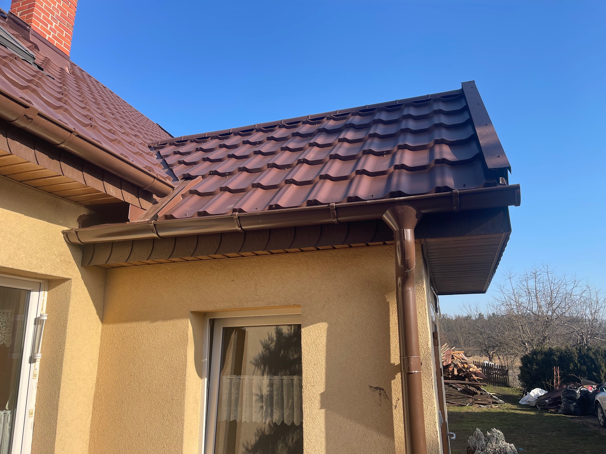 Brązowy dach z blachodachówki trapezowej i orynnowanie na tle błękitnego nieba. Widoczny fragment elewacji budynku z oknami i komin z cegły.