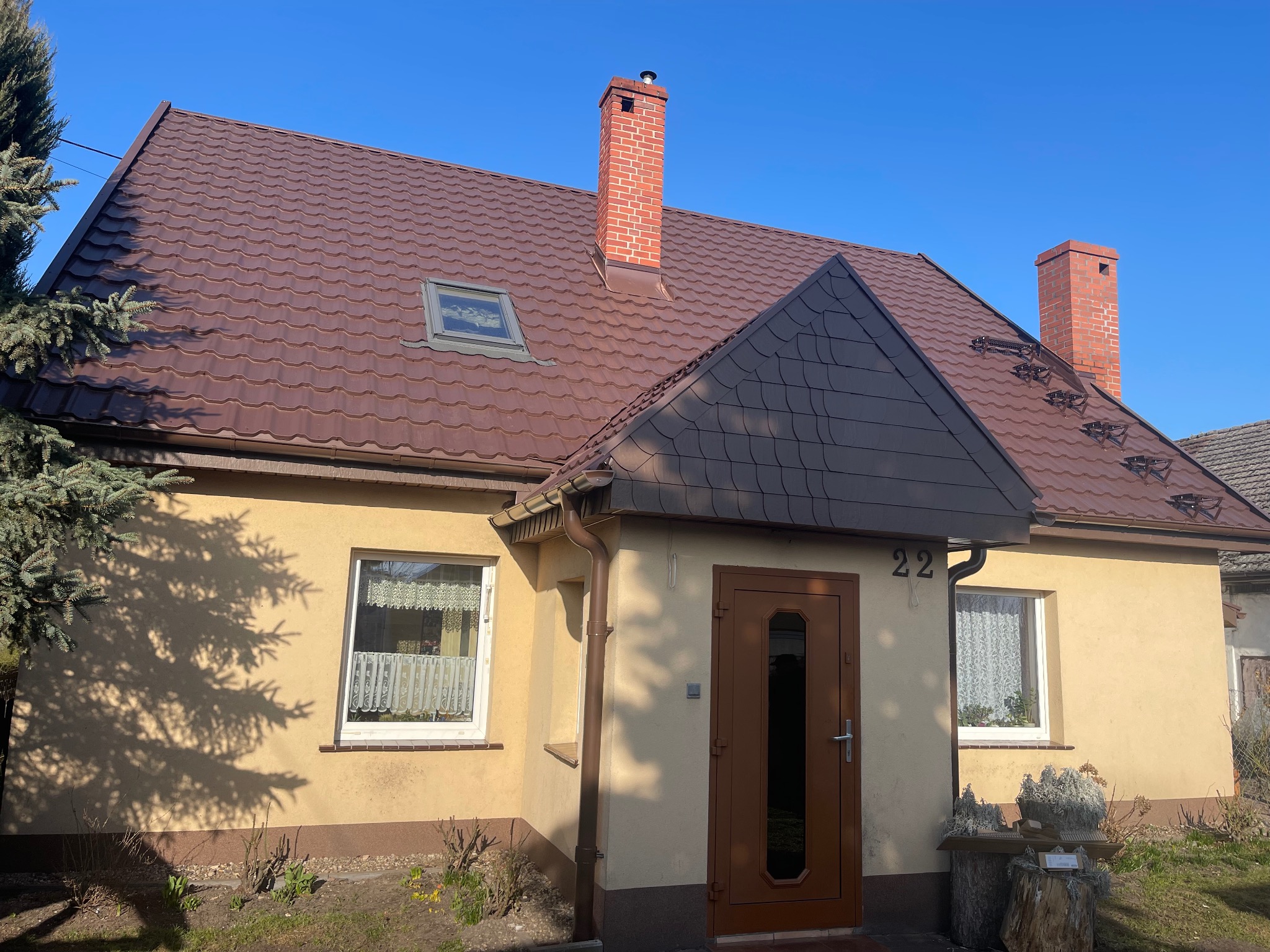 Dom jednorodzinny z brązowym dachem z blachodachówki, ceglanym kominem i numerem 22 nad wejściem, sfotografowany w słoneczny dzień.
