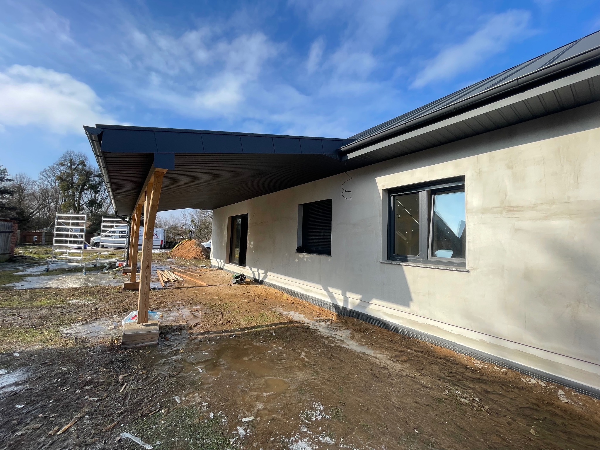 Nowoczesny dom jednorodzinny w trakcie budowy, z widocznym zadaszeniem tarasu wspartym na drewnianych słupach i fragmentem dachu w kolorze grafitowym.