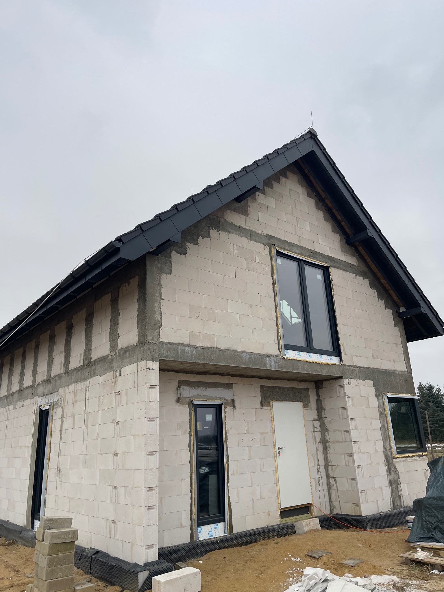 Surowy stan domu jednorodzinnego z czarną dachówką, widoczne okna i drzwi. Budynek w trakcie budowy, ściany z betonu komórkowego, szara, pochmurna pogoda.