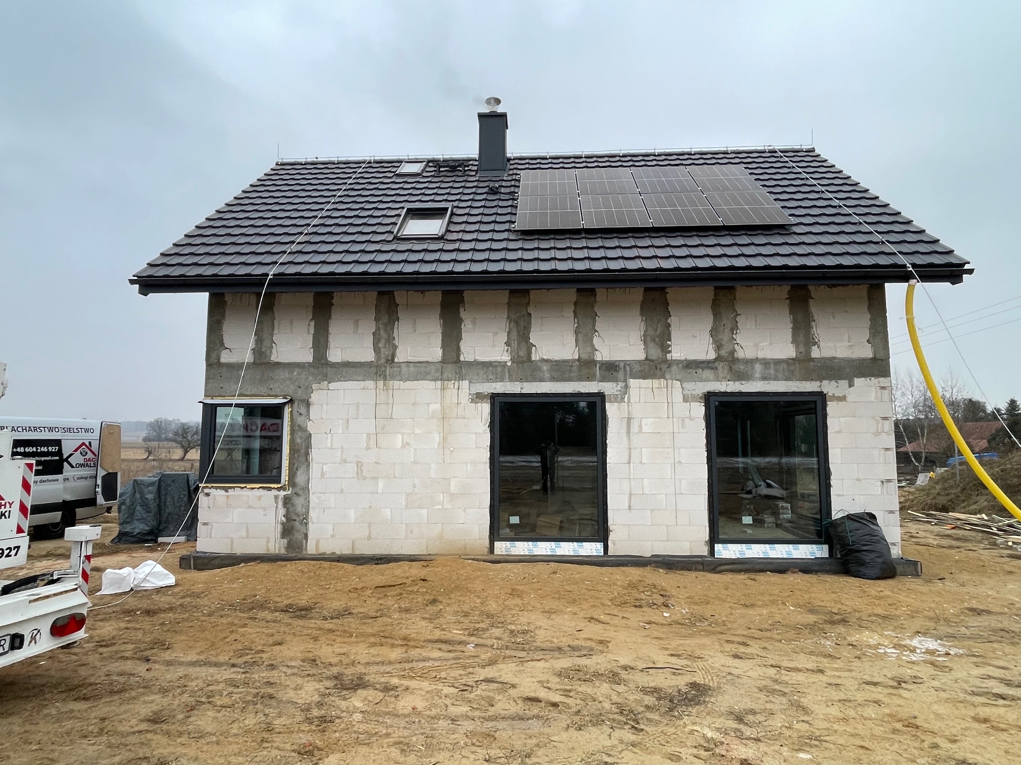 Dom w trakcie budowy z czarną dachówką, oknami i panelami słonecznymi na dachu. Widoczne niedokończone ściany z betonu komórkowego i elementy rusztowania.