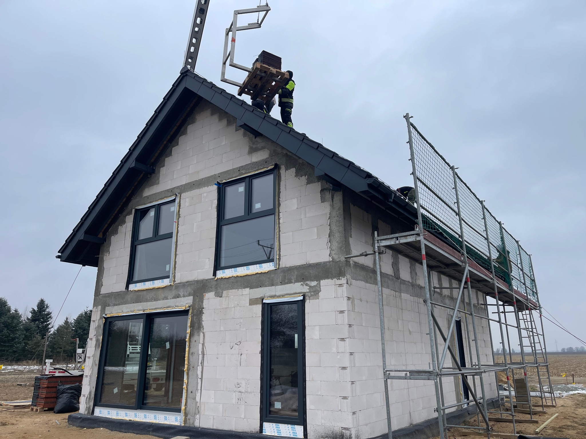 Montaż czarnej dachówki na nowym domu z białego bloczka. Na dachu pracownicy w odblaskowych ubraniach. Widoczne rusztowanie i podnośnik do transportu materiałów.