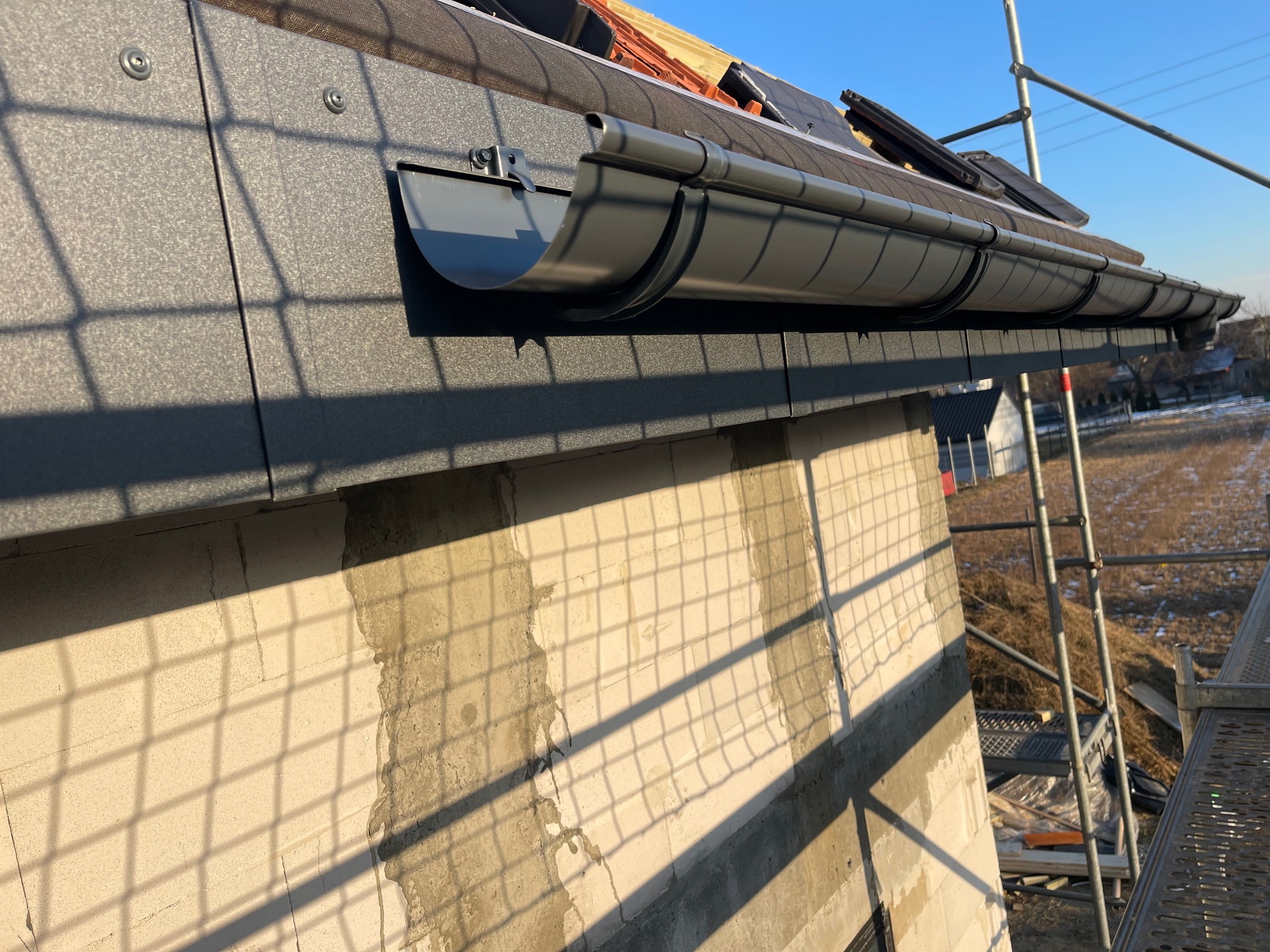 Szary system rynnowy zamontowany na budynku w trakcie budowy, widoczne rusztowania i fragment dachu z dachówką. Słoneczny dzień, cień kratownicy na ścianie.