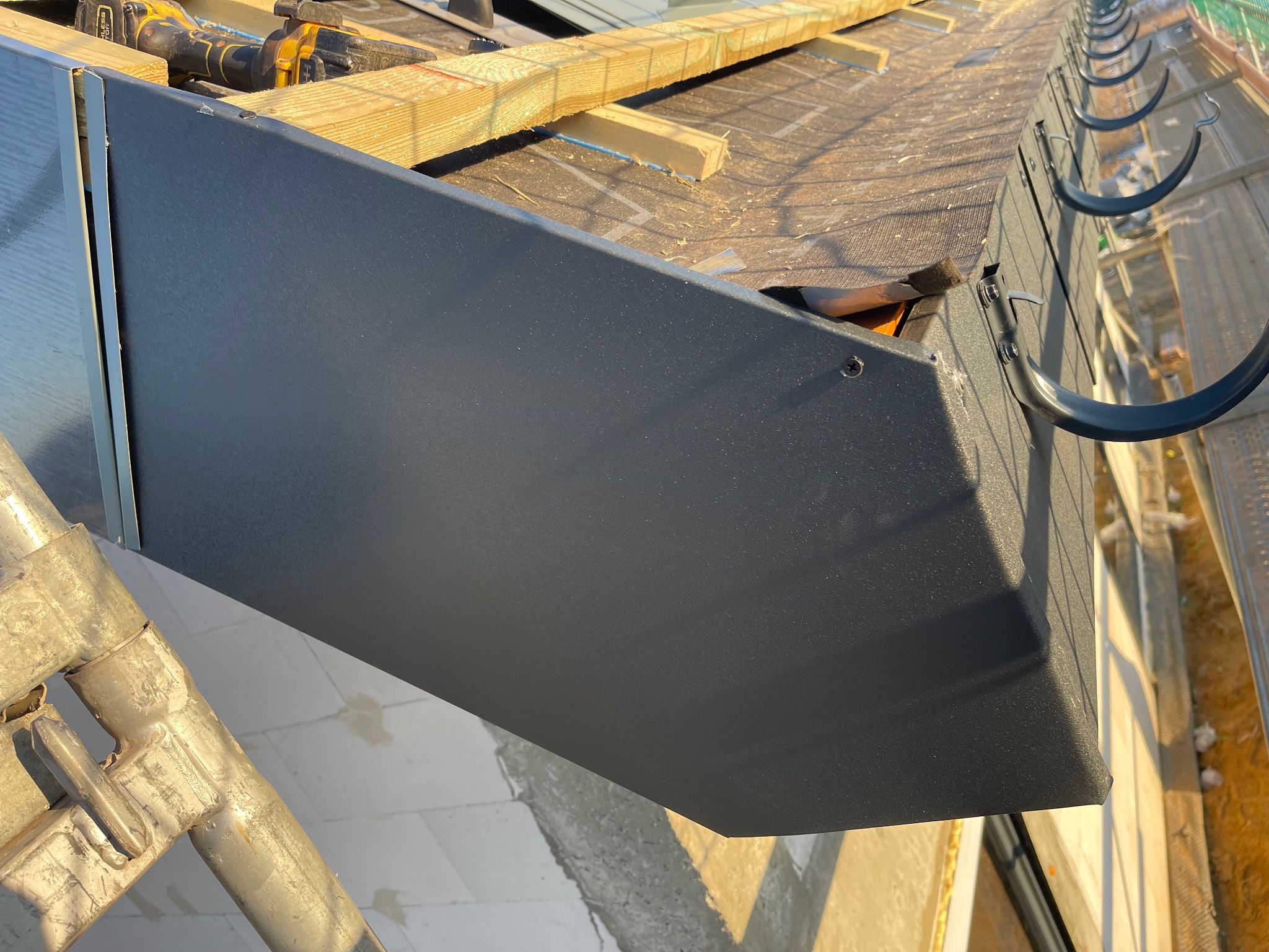 Montaż obróbki blacharskiej dachu z czarnymi rynnami, widok z rusztowania. Na dachu widoczne narzędzia i drewniane elementy konstrukcyjne.