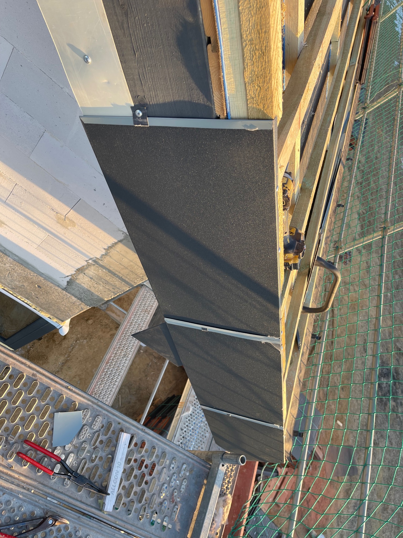 Fragment konstrukcji dachu z widoczną membraną, elementami drewnianymi i metalowymi, zabezpieczony siatką. Na rusztowaniu leżą narzędzia dekarskie.