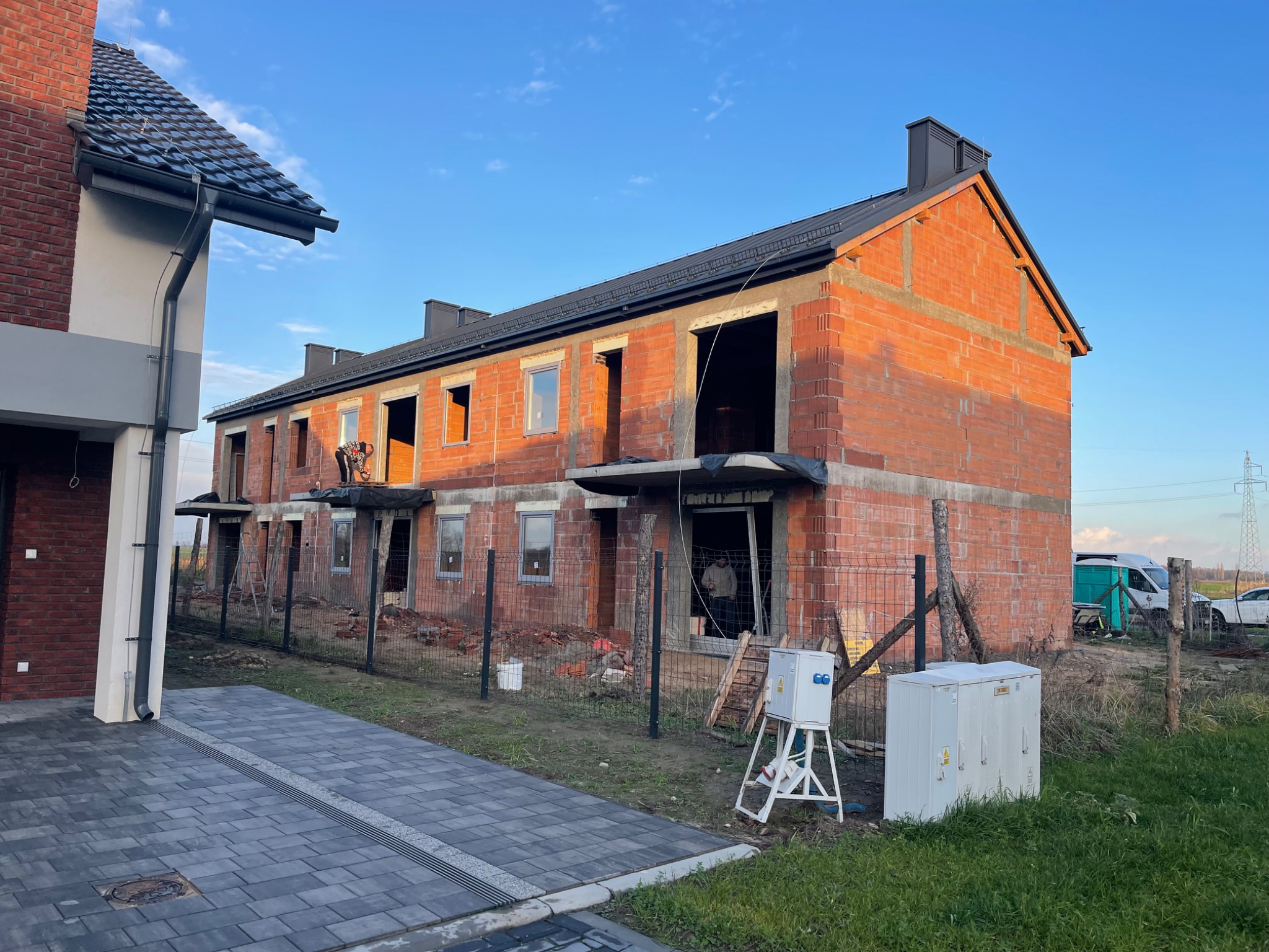 Surowy stan szeregowca z cegły, z czarną dachówką i kominami, w trakcie budowy. Widoczny pracownik na dachu. Ujęcie z zewnątrz, w słoneczny dzień.