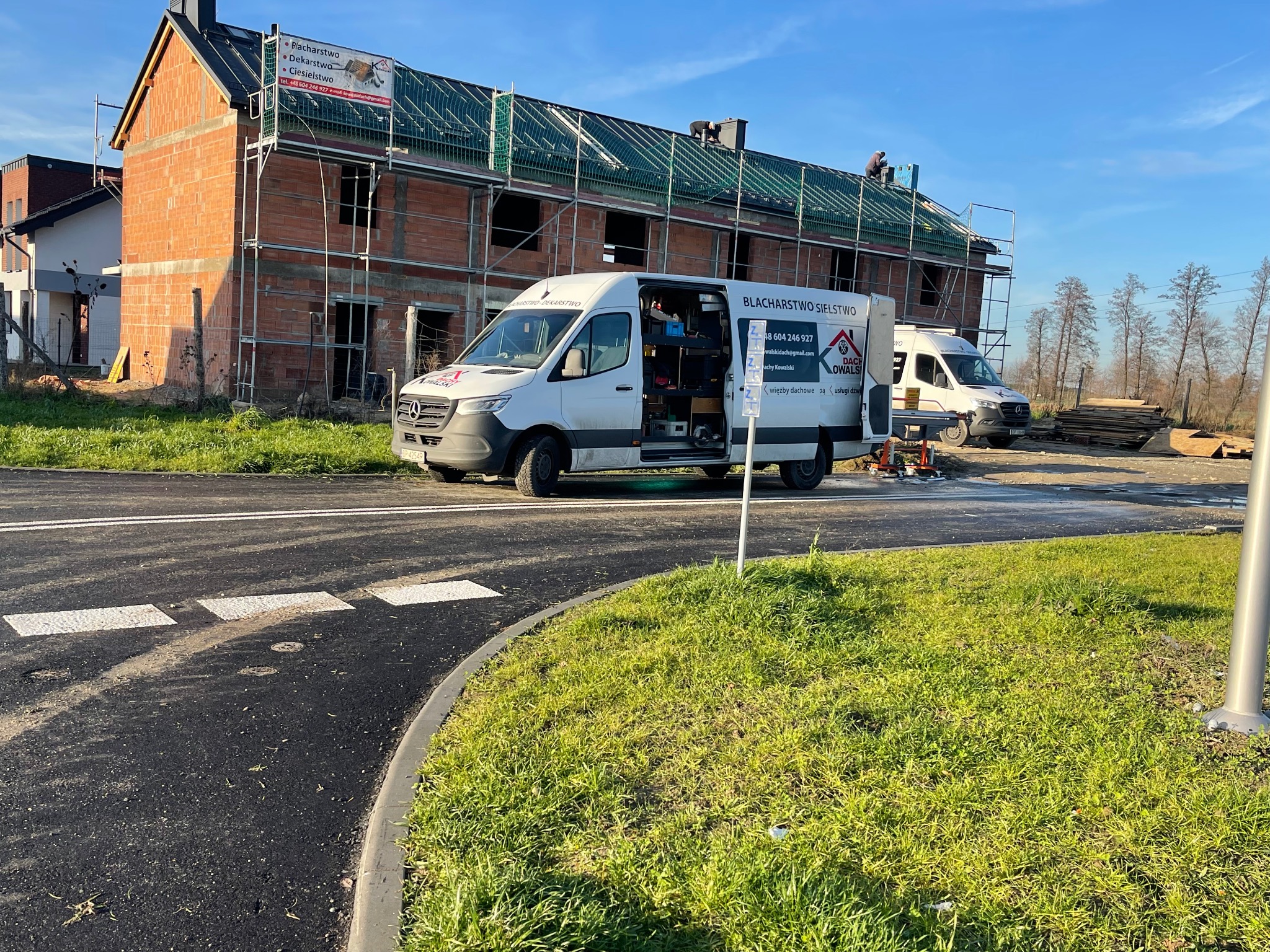 Budowa domu z częściowo wykonanym dachem, widoczni dekarze na dachu, na pierwszym planie samochód dostawczy z reklamą firmy dekarskiej.