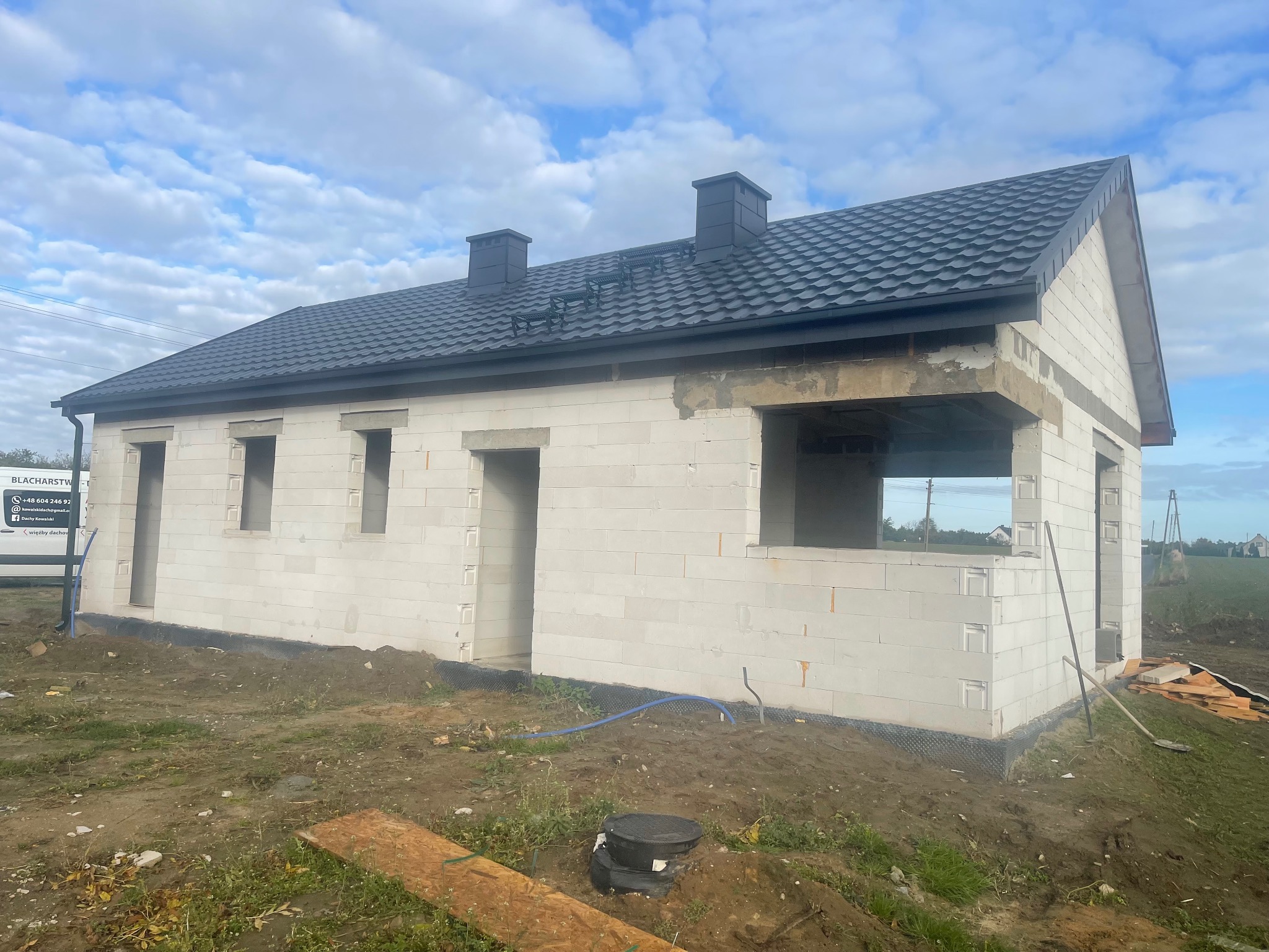 Niedokończony dom z ciemnym dachem z blachy na tle błękitnego nieba. Widoczne kominy i elementy konstrukcyjne. Na pierwszym planie ziemia i resztki materiałów budowlanych.