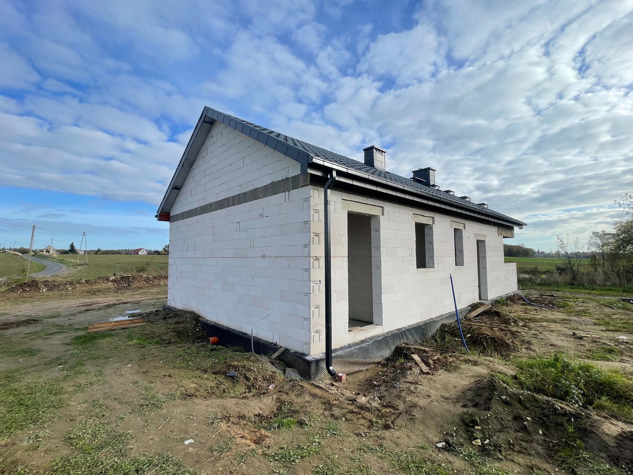 Niedokończony dom z białego pustaka z czarnym dachem i rynnami na tle błękitnego nieba z chmurami. Widoczne okna i drzwi w stanie surowym.