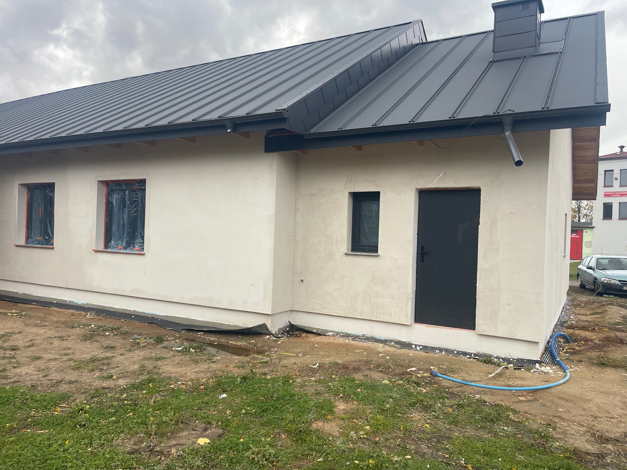 Nowoczesny dom jednorodzinny z ciemnoszarą blachodachówką i świeżo otynkowaną elewacją. Okna zabezpieczone folią, widoczny fragment ogrodu i niebieski wąż.