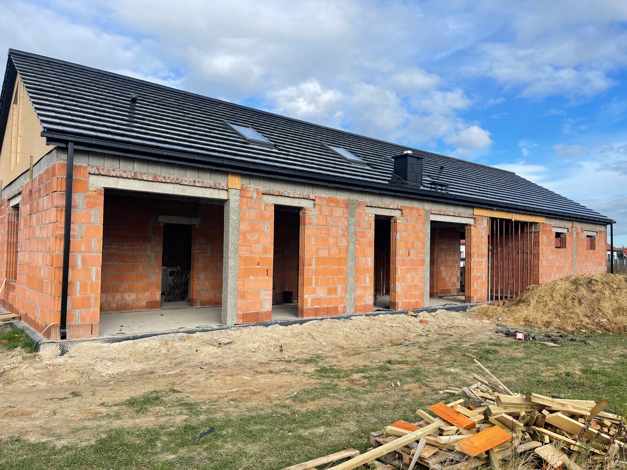 Dom w trakcie budowy z czarnym dachem i ceglanymi ścianami. Widoczne okna dachowe i komin. Na pierwszym planie sterta drewna i piasku.