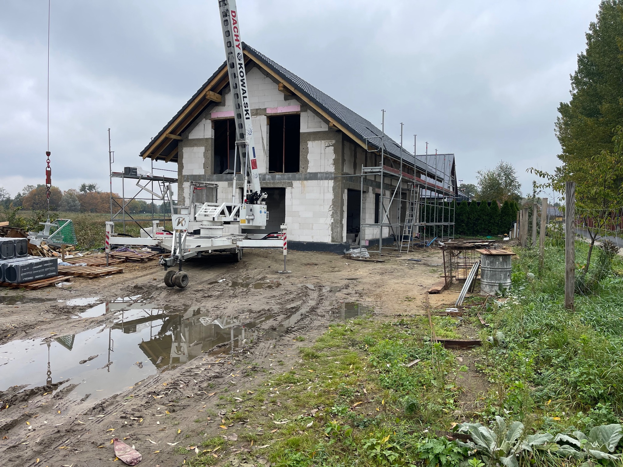 Budowa domu jednorodzinnego w trakcie prac. Widoczny dźwig, rusztowania i częściowo położone pokrycie dachu. Teren budowy błotnisty po deszczu.