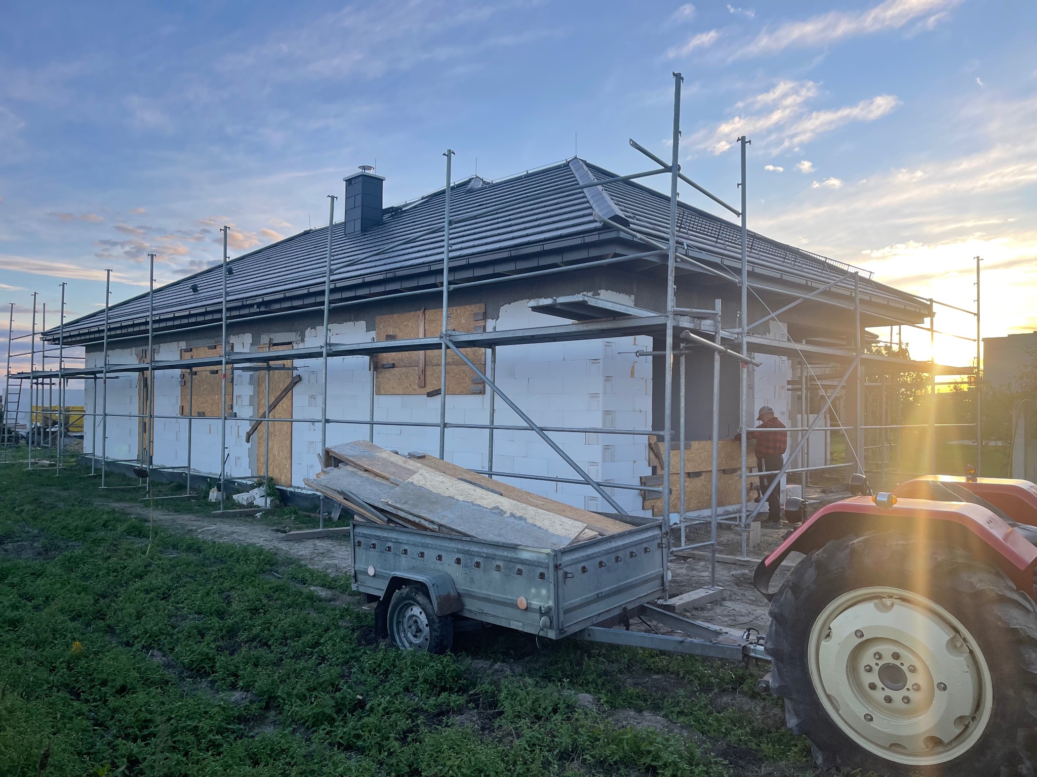 Dom w trakcie budowy z rusztowaniem i przyczepką. Widoczny fragment traktora. Dach pokryty szarą dachówką. Prace budowlane w toku, ściany ocieplone styropianem.