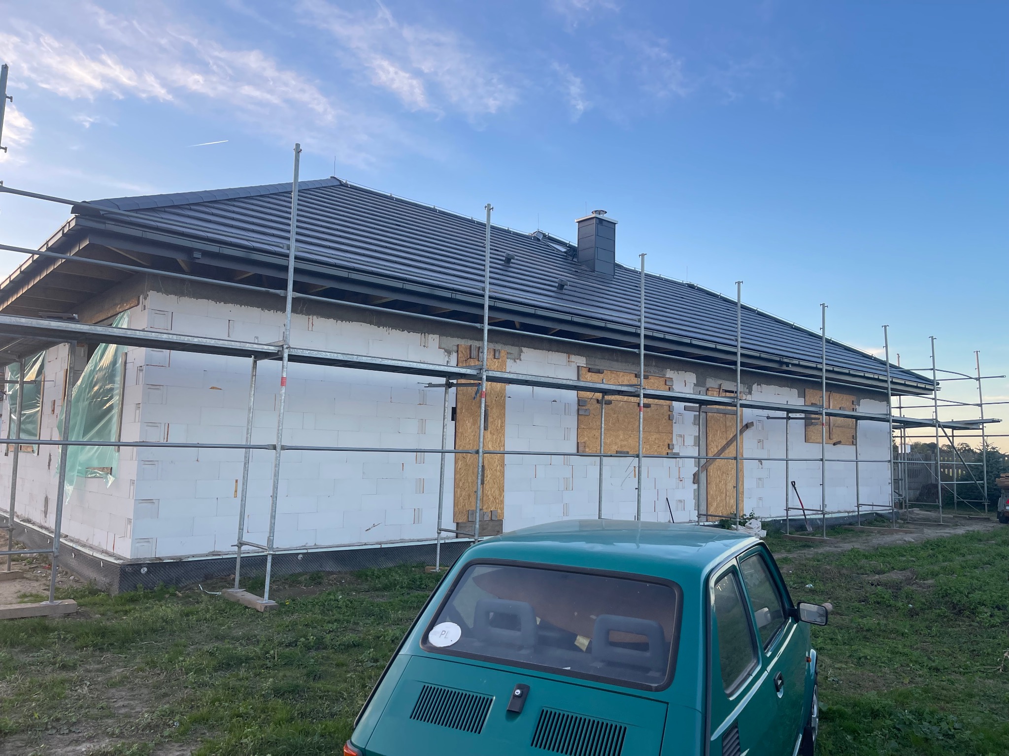 Dom w trakcie budowy z szarym dachem, otoczony rusztowaniami, z zielonym samochodem na pierwszym planie i błękitnym niebem w tle.