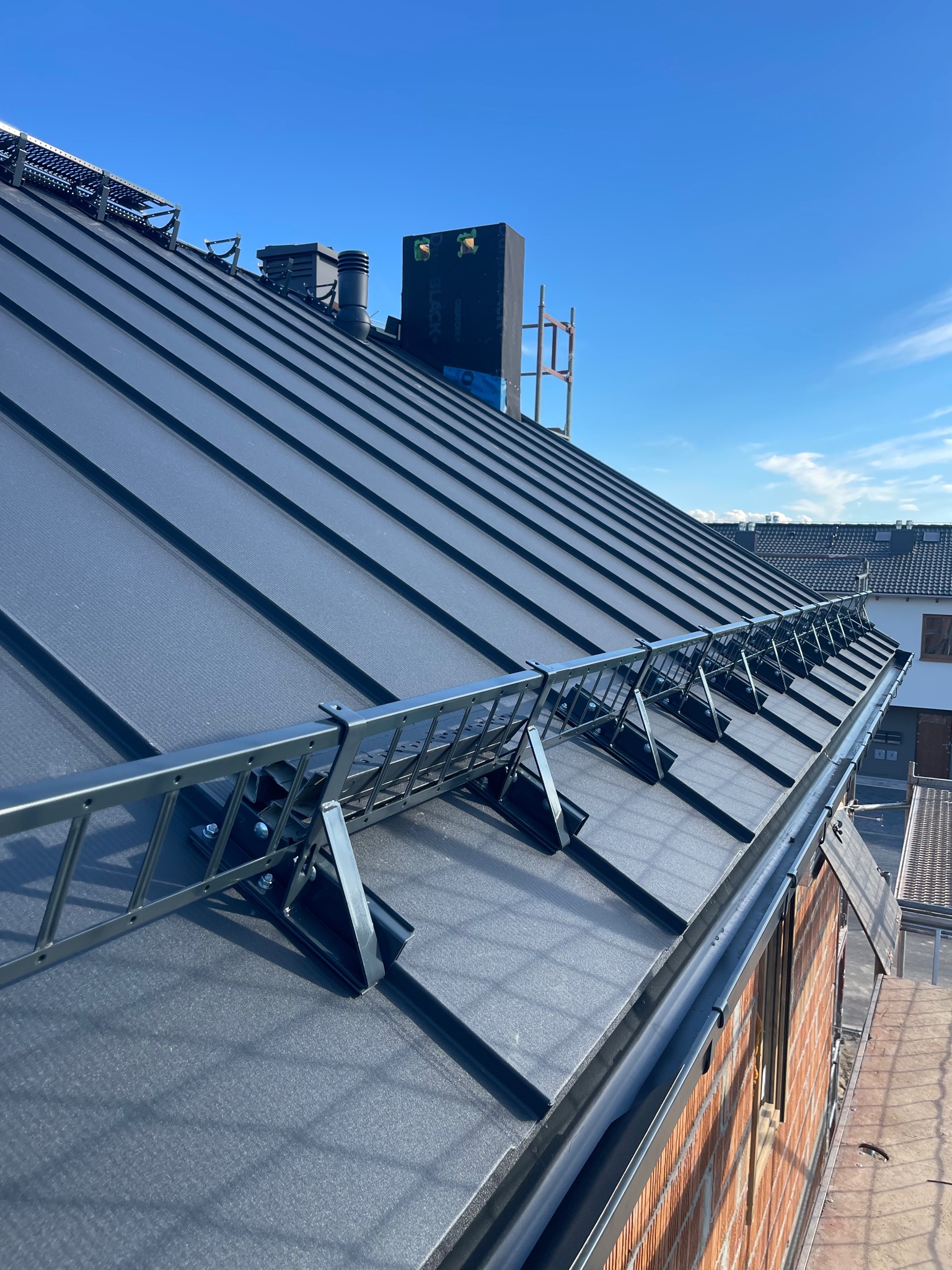 Czarny, blaszany dach z zamontowanymi zabezpieczeniami przeciwśniegowymi i kominami wentylacyjnymi na tle błękitnego nieba.