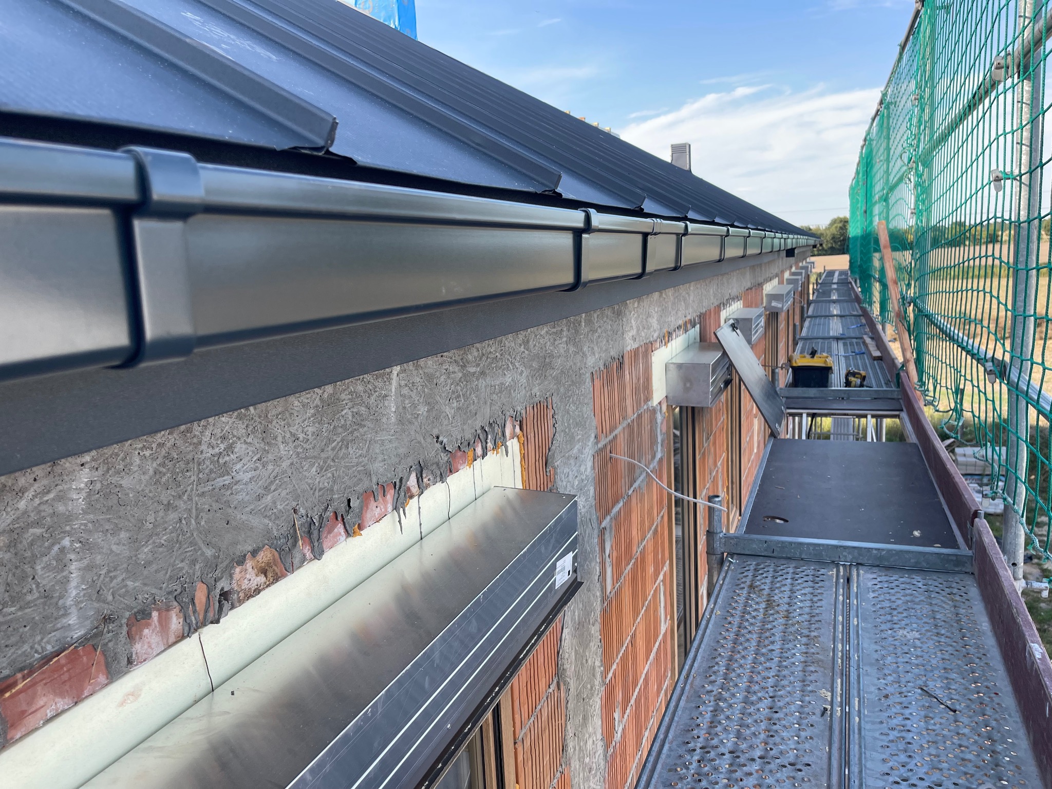 Zamontowane, czarne orynnowanie dachu na budynku z cegły, widoczne z rusztowania. Fragment dachu z blachy. Siatka zabezpieczająca na rusztowaniu.