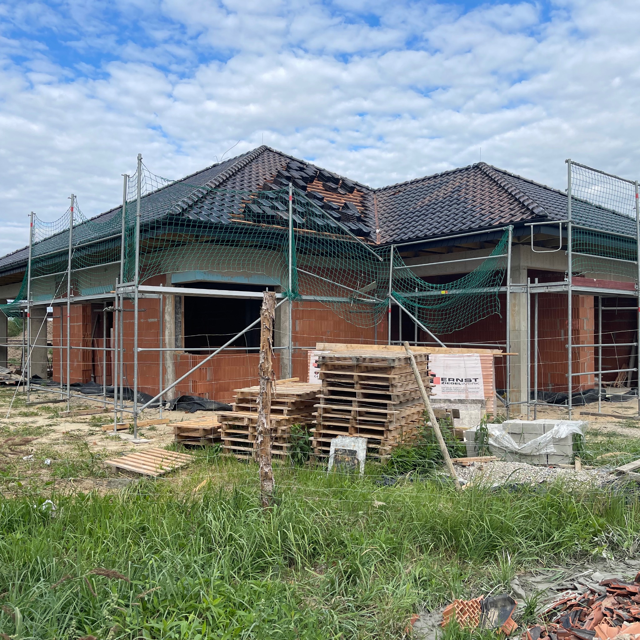 Budowa domu jednorodzinnego w trakcie: widoczne ściany z cegły, rusztowania, palety z materiałami budowlanymi i częściowo zdjęte dachówki na fragmencie dachu.